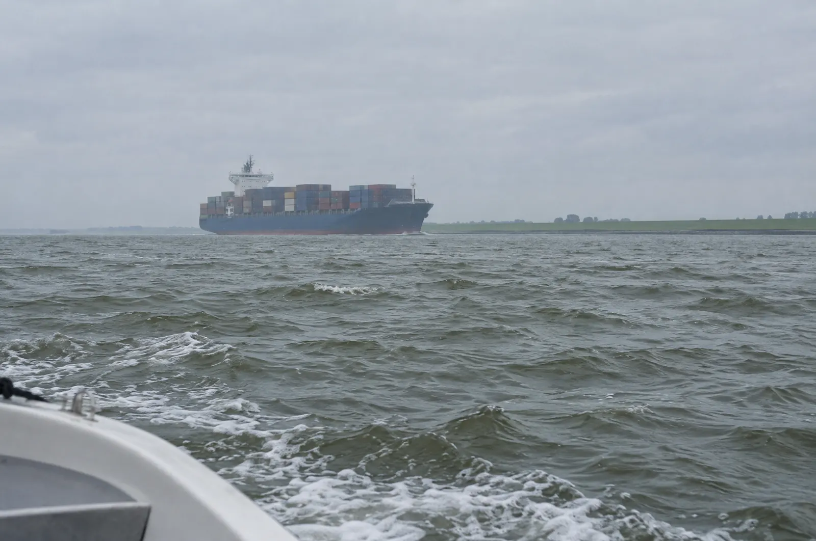 Containerschiff auf einem breiten Fluss an einem grauen Tag, gesehen vom Bug eines kleinen Sportboots, am anderen Ufer ein flacher grüner Deich