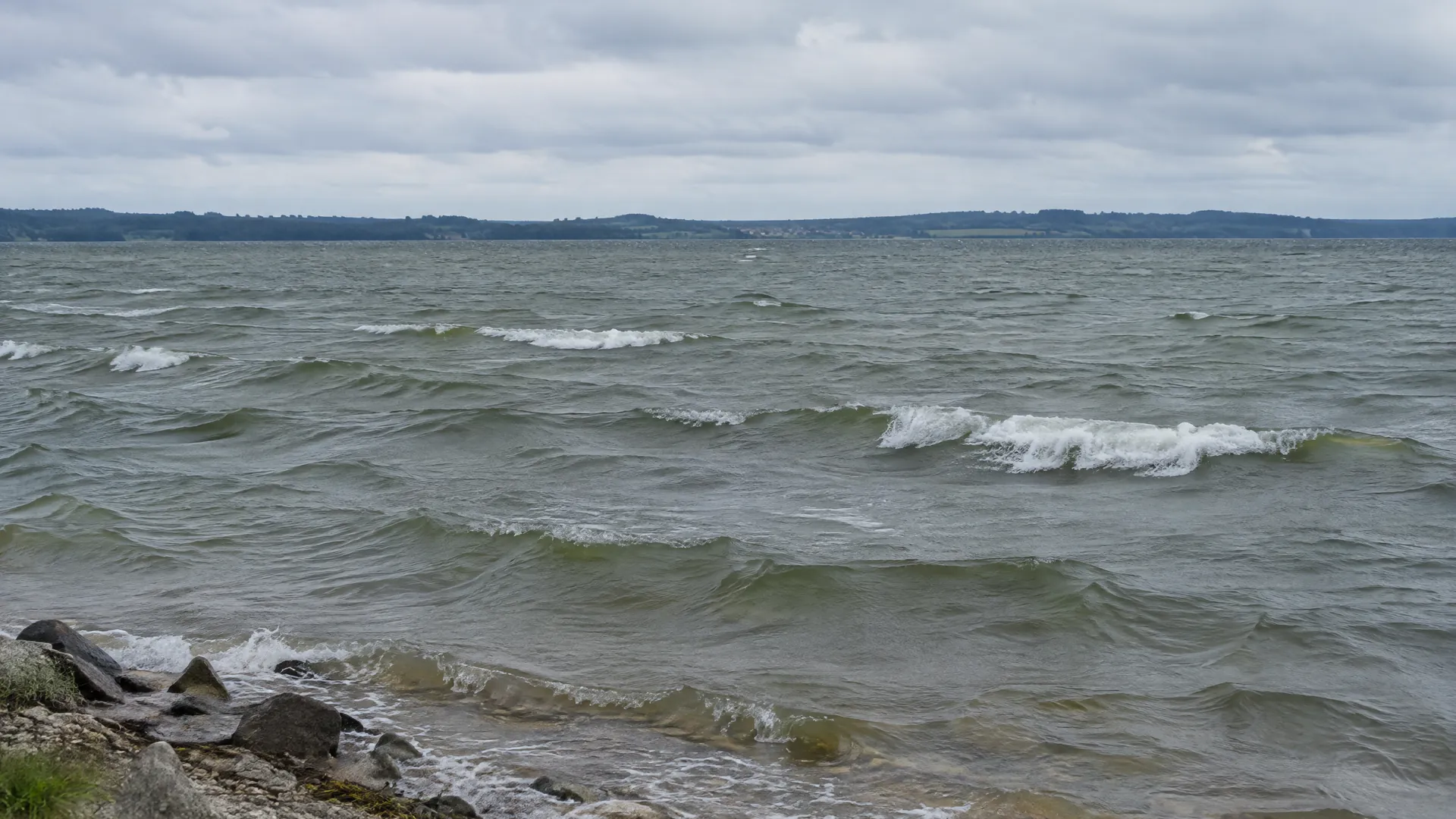 Kurze Windwellen mit einzelnen weißen Kämmen auf einem See unter bedecktem Himmel