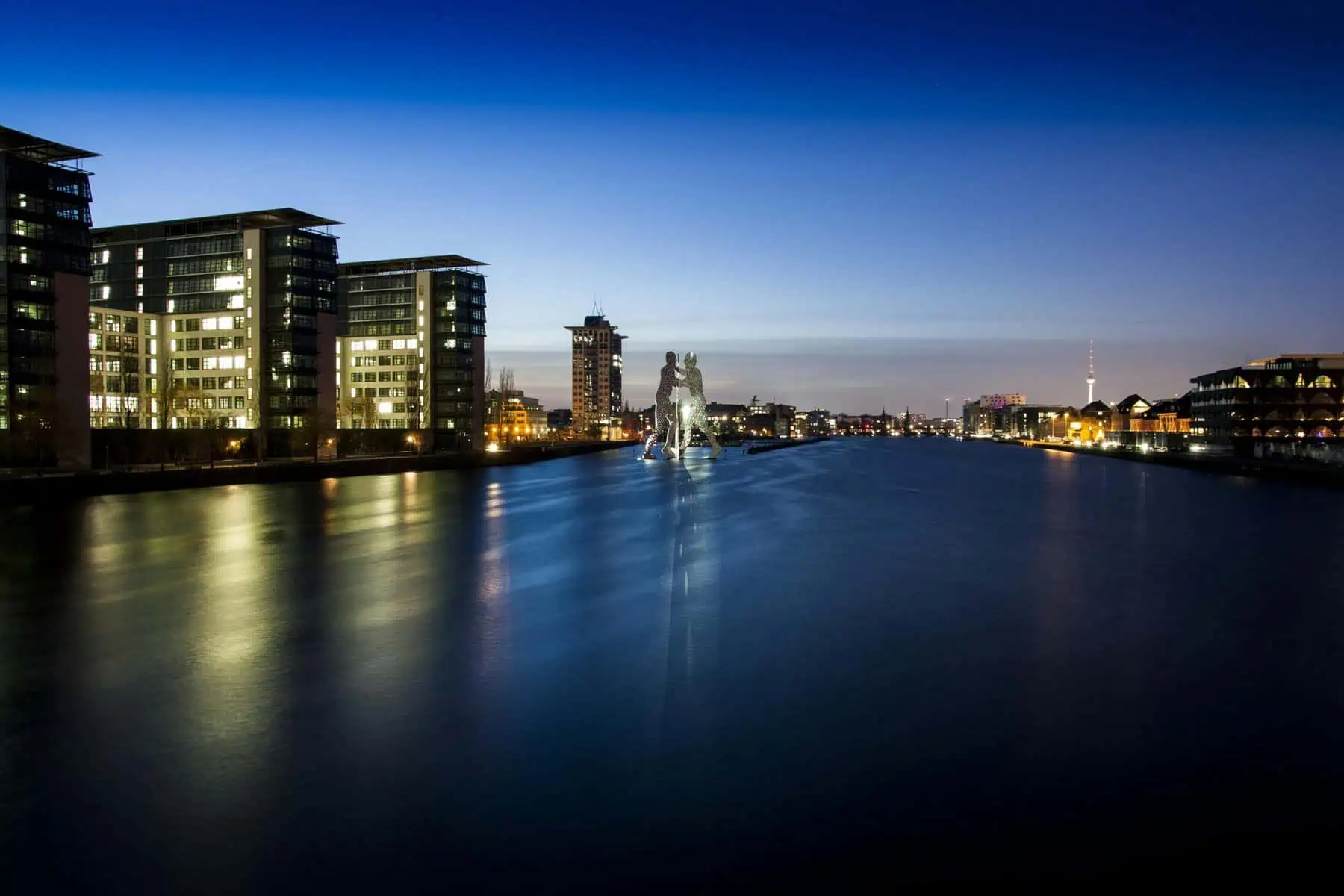 Spree in Berlin zur blauen Stunde: die Skulptur Molecule Man steht im Fluss, links beleuchtete Bürohäuser am Ufer, am Horizont der Fernsehturm