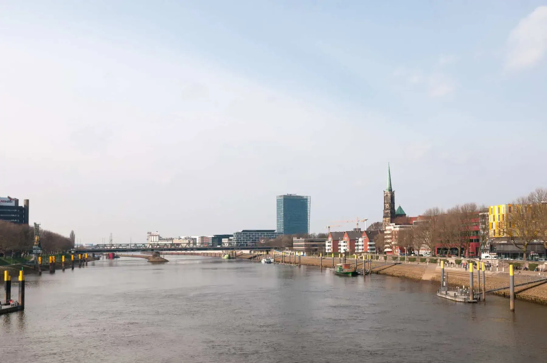 Die Weser in Bremen im Morgenlicht: rechts die Uferpromenade mit Anlegern und Dalben, im Hintergrund eine Brücke, ein Hochhaus und ein Kirchturm
