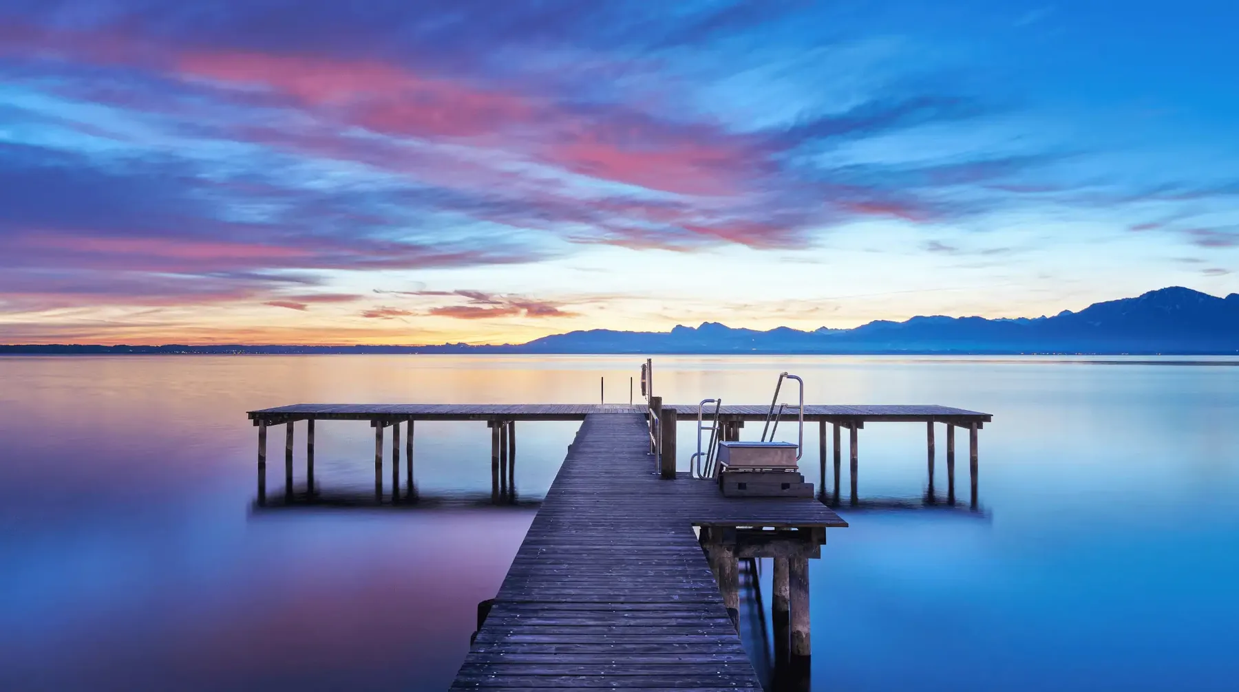 Holzsteg mit Badeleiter im stillen Wasser eines Sees bei Abendrot, am Horizont eine Bergkette