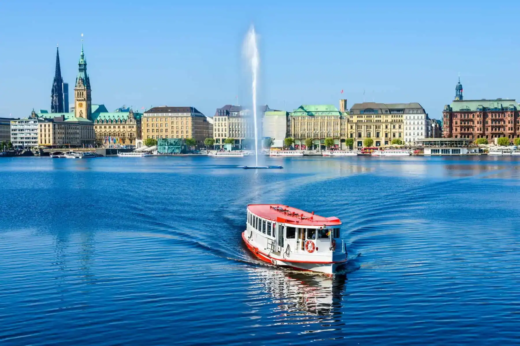 Binnenalster in Hamburg bei Sonne: ein Alsterdampfer fährt auf die Kamera zu, dahinter die Alsterfontäne, das Rathaus und die Häuserzeile am Jungfernstieg