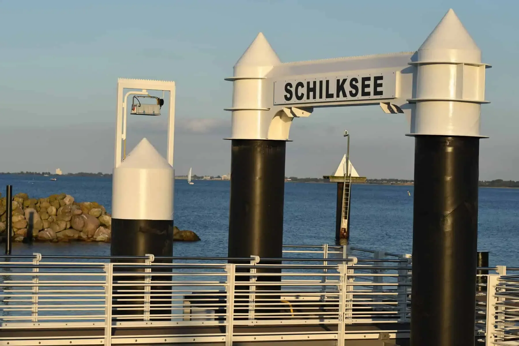 Fähranleger Kiel-Schilksee im Abendlicht: weißes Portal mit der Aufschrift „Schilksee“, dahinter die Kieler Förde mit einem Segelboot
