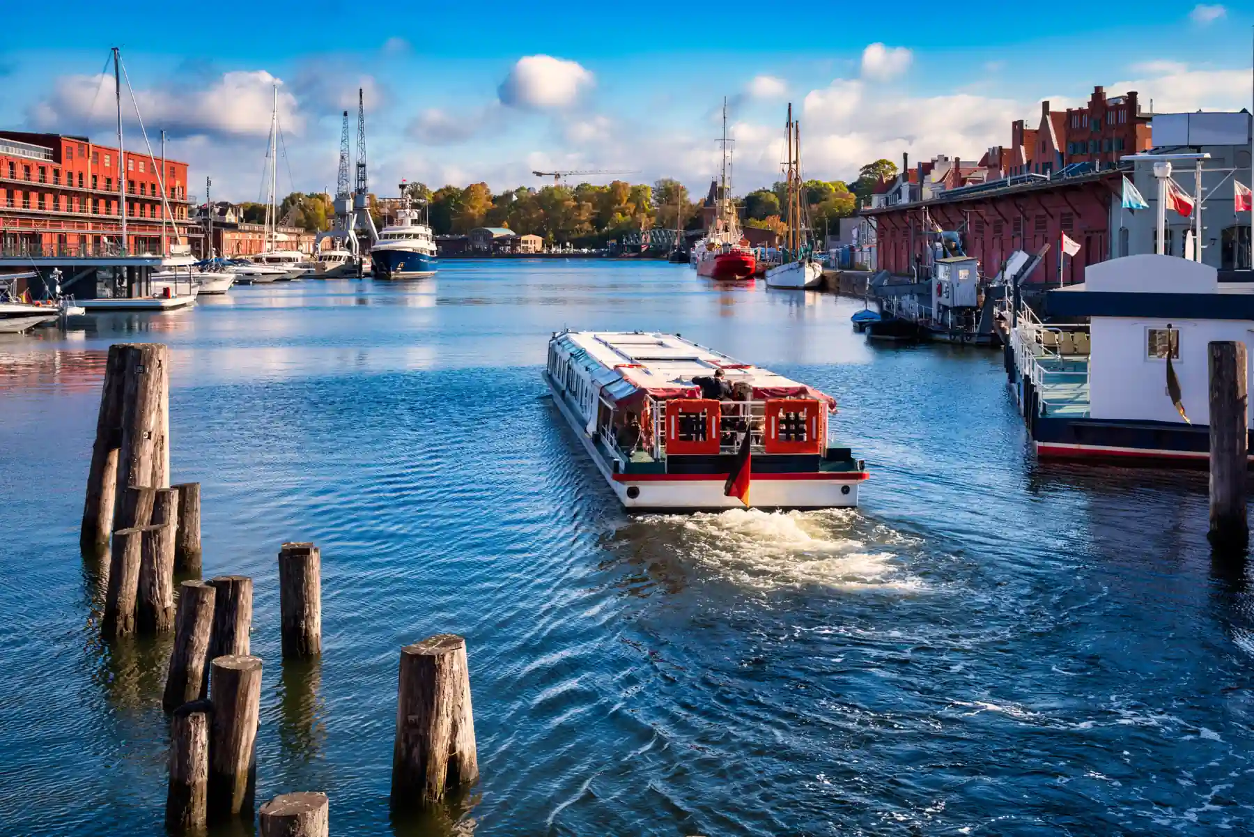 Hafen an der Trave in Lübeck bei Sonne: ein Fahrgastschiff fährt zwischen Holzdalben vom Betrachter weg, links ein roter Backsteinbau mit Sportbooten und Hafenkränen, rechts Traditionsschiffe vor alten Lagerhäusern