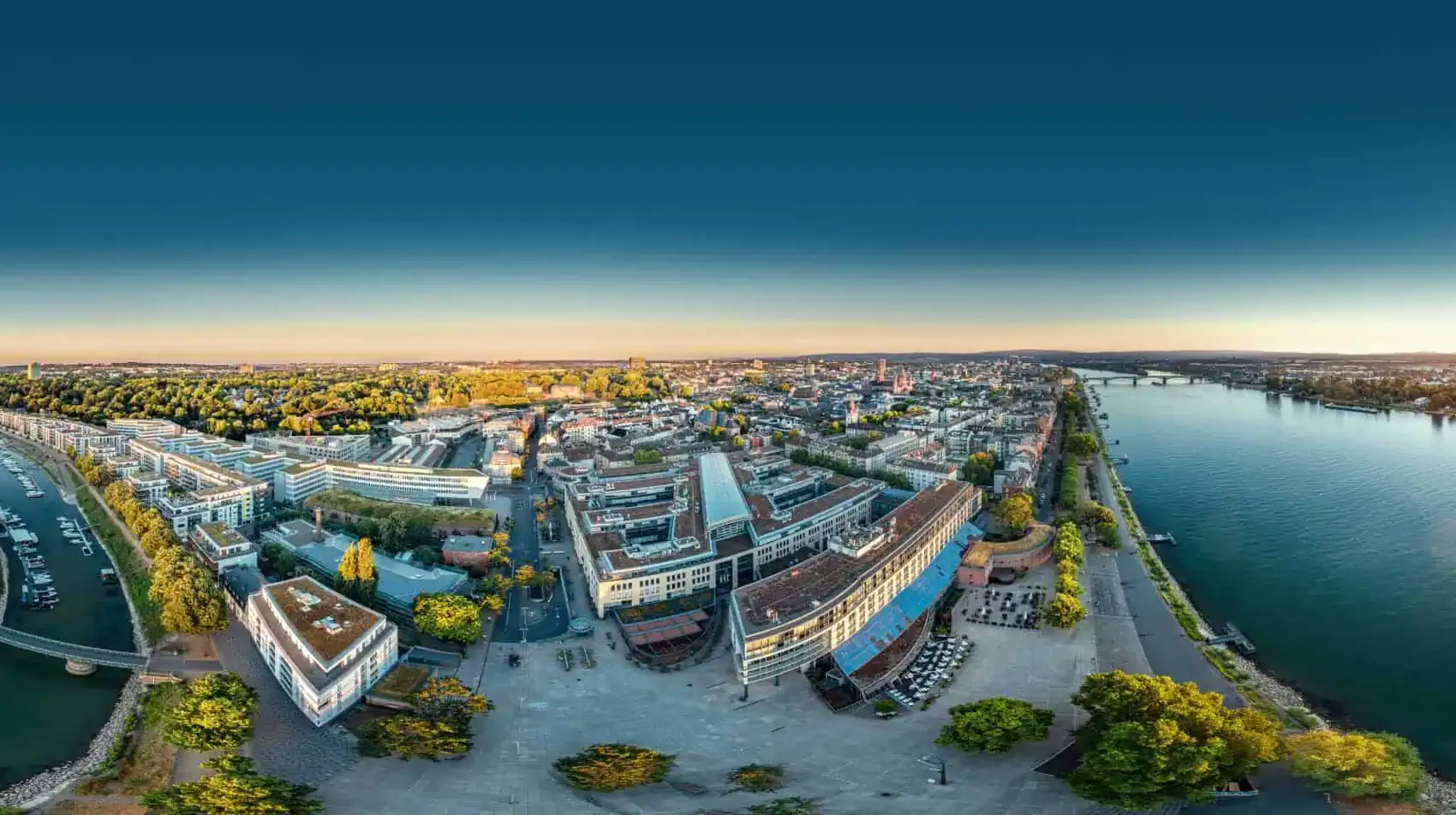 Luftbild von Mainz im Abendlicht als Weitwinkel-Panorama: rechts der Rhein mit Uferpromenade und Brücke, links ein Hafenbecken mit Sportbooten, dazwischen Häuserblöcke und ein großer Platz