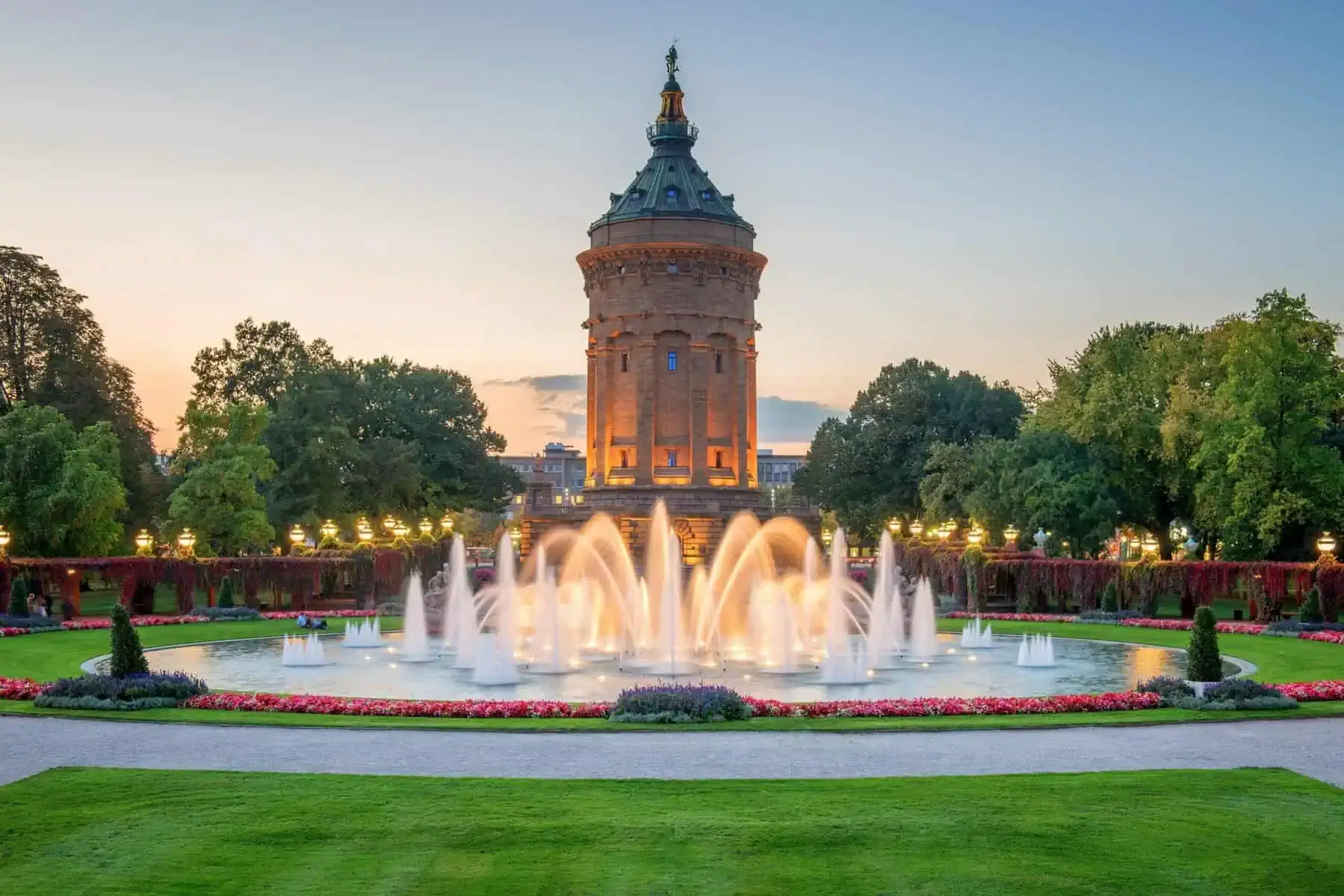 Der Mannheimer Wasserturm in der Abenddämmerung, davor beleuchtete Wasserfontänen in einem runden Becken, Blumenbeete und Rasen
