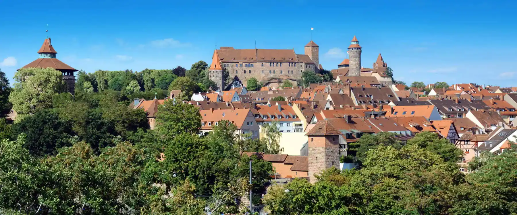 Blick über die Dächer der Nürnberger Altstadt zur Kaiserburg mit dem runden Sinwellturm, davor Bäume, darüber blauer Himmel