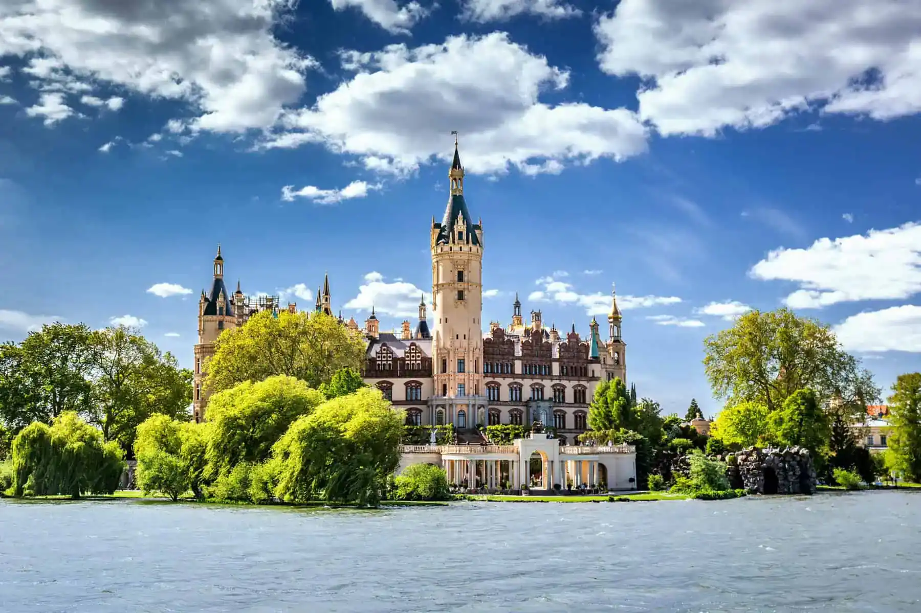 Das Schweriner Schloss bei Sonne und weißen Wolken, davor Bäume, ein Säulengang am Ufer und im Vordergrund Wasser