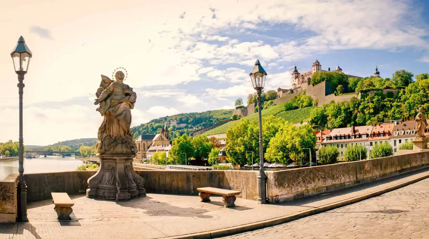 Auf der Alten Mainbrücke in Würzburg: eine Heiligenstatue und Laternen, links der Main, dahinter die Festung Marienberg über Weinbergen